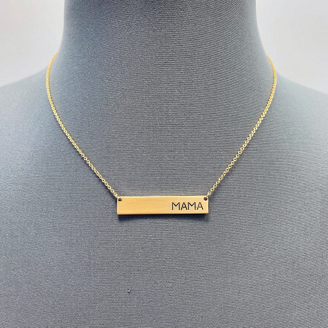 A collection of four necklaces with metal bars engraved with the word 'MAMA', displayed on a mannequin. Two necklaces are gold-colored, and two are silver-colored.