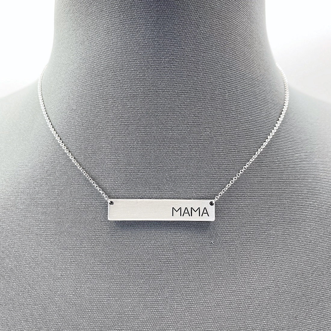 A collection of four necklaces with metal bars engraved with the word 'MAMA', displayed on a mannequin. Two necklaces are gold-colored, and two are silver-colored.