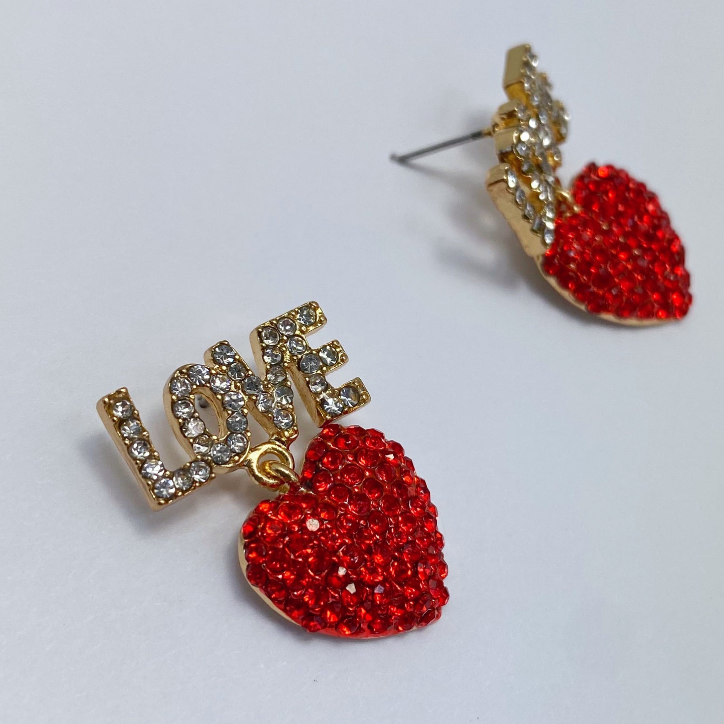 A set of red heart-shaped earrings with the word 'LOVE' written in rhinestones, displayed on a mannequin head from different angles.