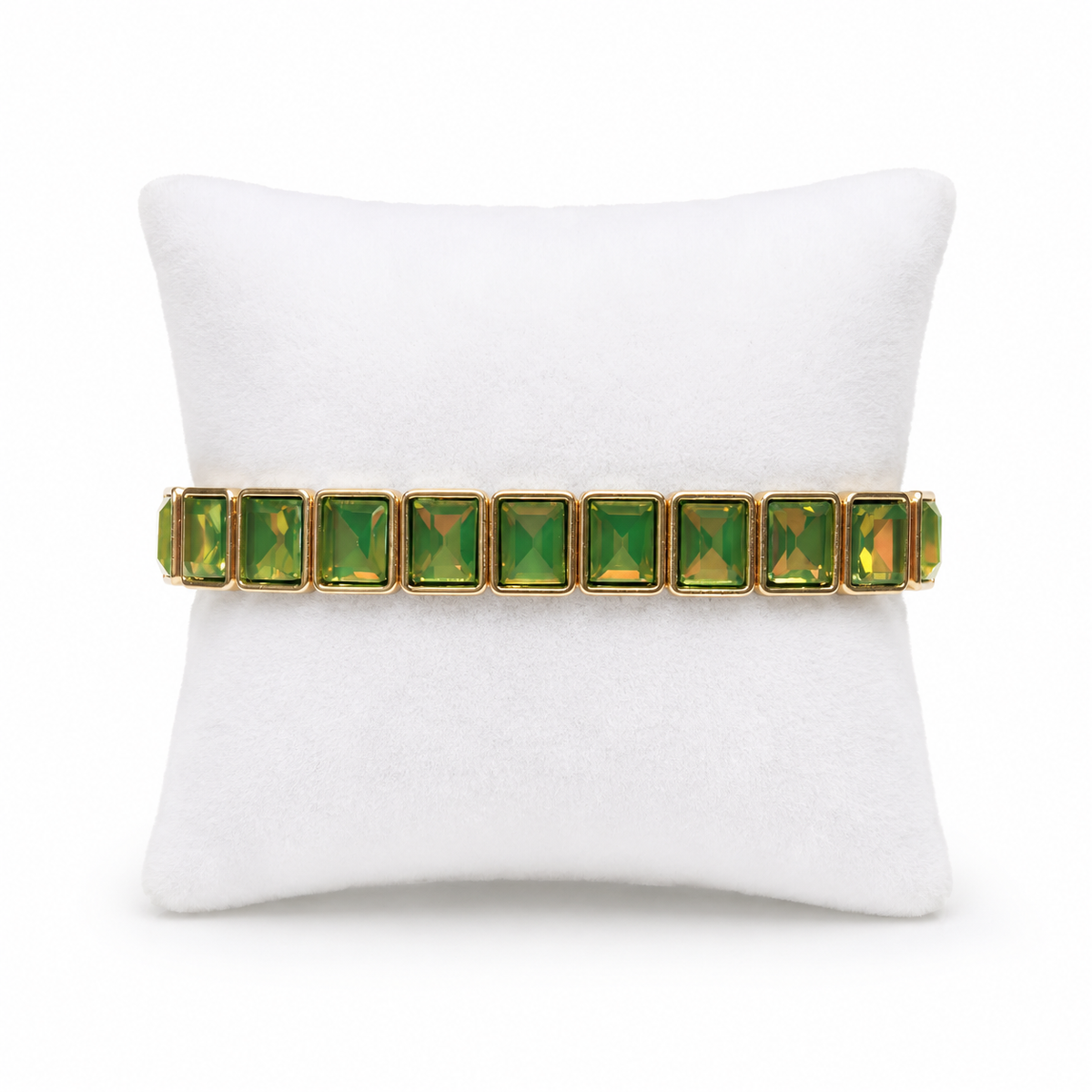 A gold-plated bracelet with a row of green rectangular faceted gemstones, displayed on a white pillow against a white background.