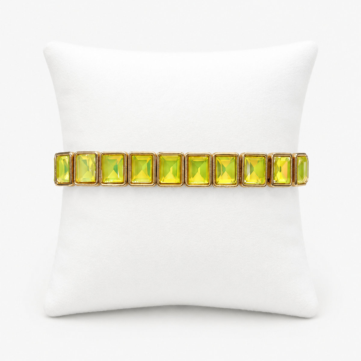 A gold-tone stretch bracelet with a row of rectangular iridescent yellow-green rhinestones, displayed on a white pillow against a light gray background.