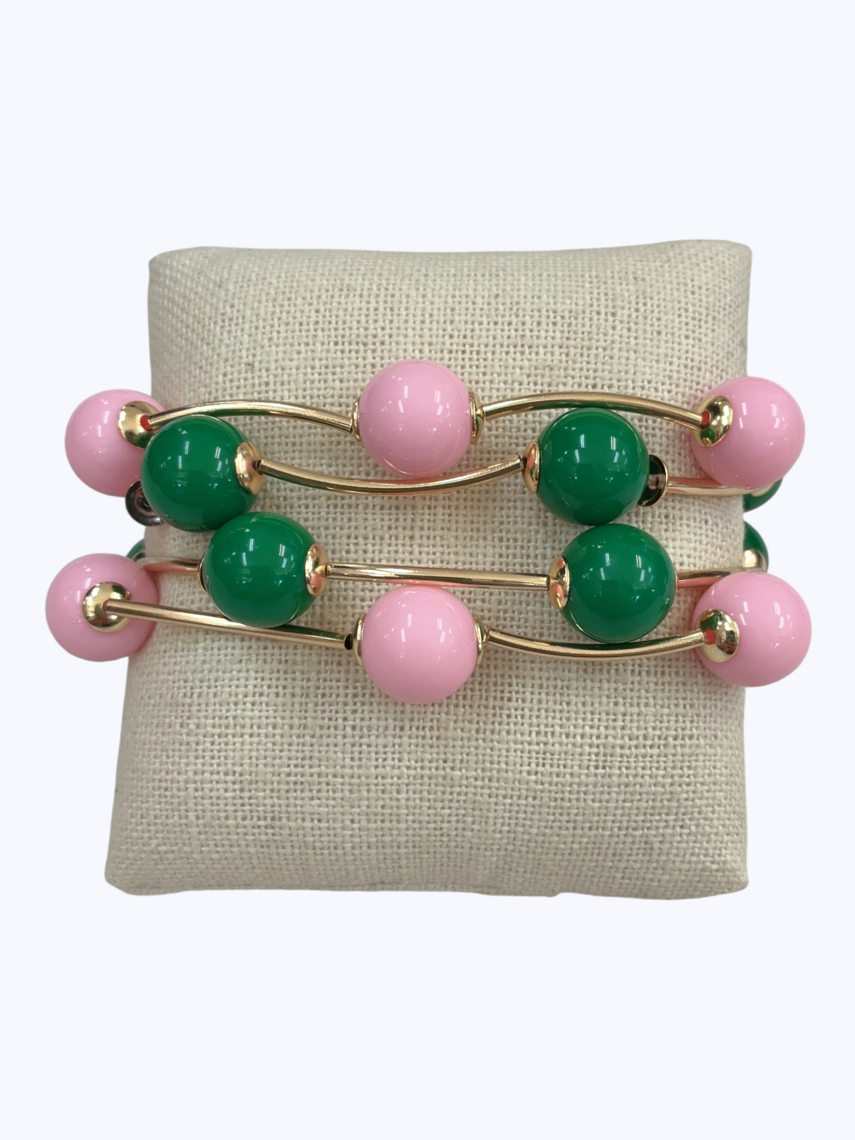 A set of three gold-plated stretch bracelets with alternating pink and green glossy beads, displayed on a beige fabric jewelry stand against a light gray background.