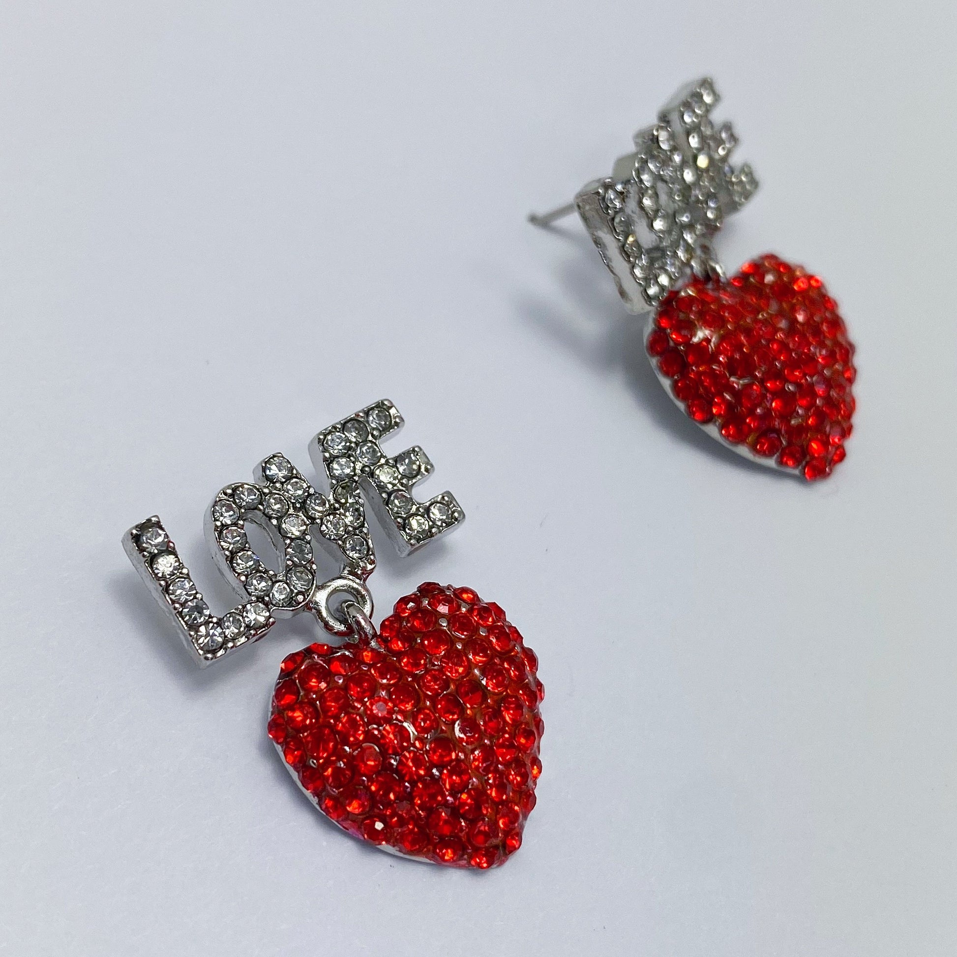 A set of red heart-shaped earrings with the word 'LOVE' written in rhinestones, displayed on a mannequin head from different angles.