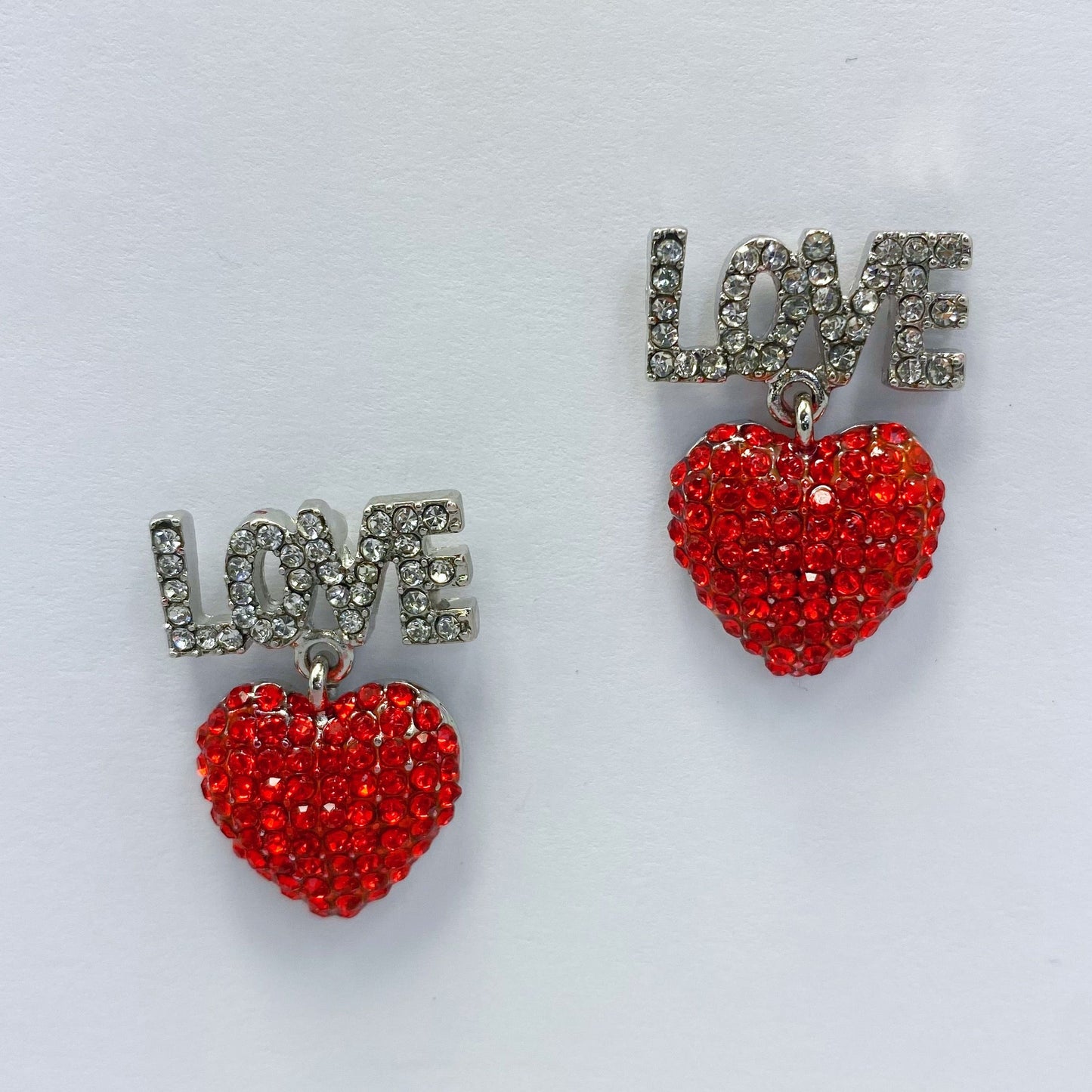 A set of red heart-shaped earrings with the word 'LOVE' written in rhinestones, displayed on a mannequin head from different angles.
